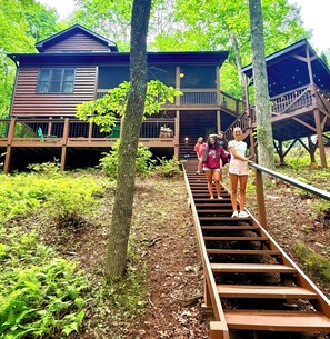 A wooden staircase leading to a cabin with a balcony, surrounded by trees and greenery.