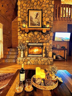 A stone fireplace with a framed picture above it, a television on the wall, and a table with candles and a bottle of wine.
