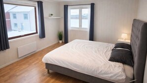 Een slaapkamer met een bed, twee ramen met gordijnen, een radiator en een kleine plant.
