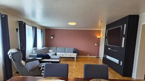 Een moderne woonkamer met een bank, stoelen, een eettafel met een kom en dienblad, en een televisie aan de muur.
