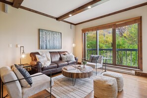 A living room with a brown leather sofa, a round coffee table, and a view of a garden through large windows. - Borders Lodge U105 2BD Beaver Creek Ski-in, Ski-out Condo (Avon)
