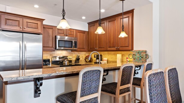Una cocina con armarios de madera, electrodomésticos de acero inoxidable, encimera de granito, taburetes de bar, iluminación colgante y salpicadero.