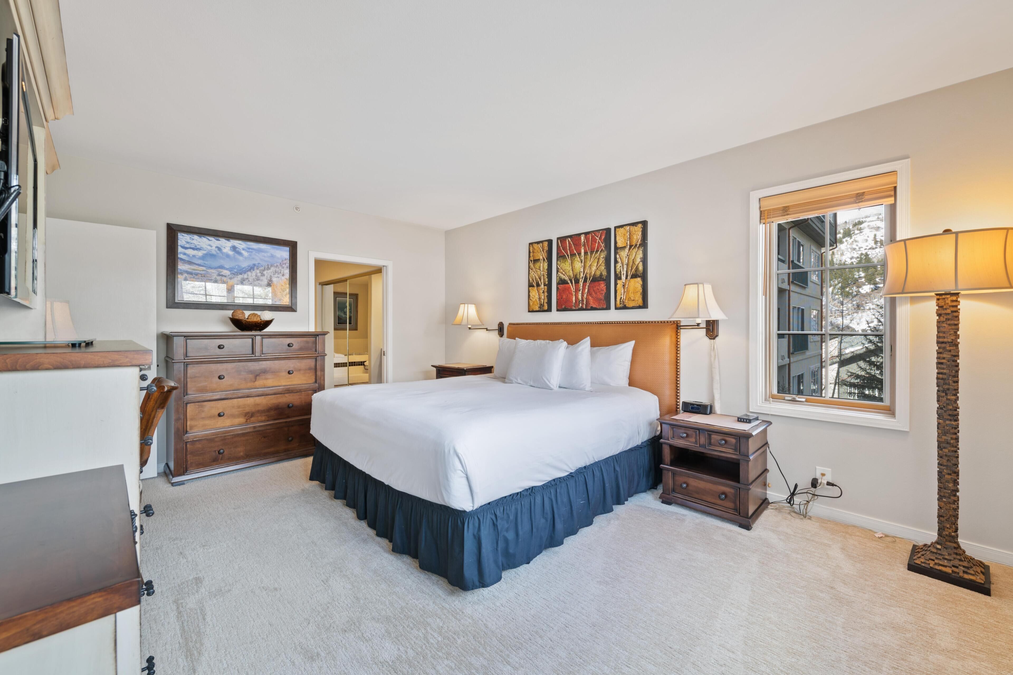 A bedroom with a large bed, a dresser, a nightstand, a lamp, and a painting on the wall.