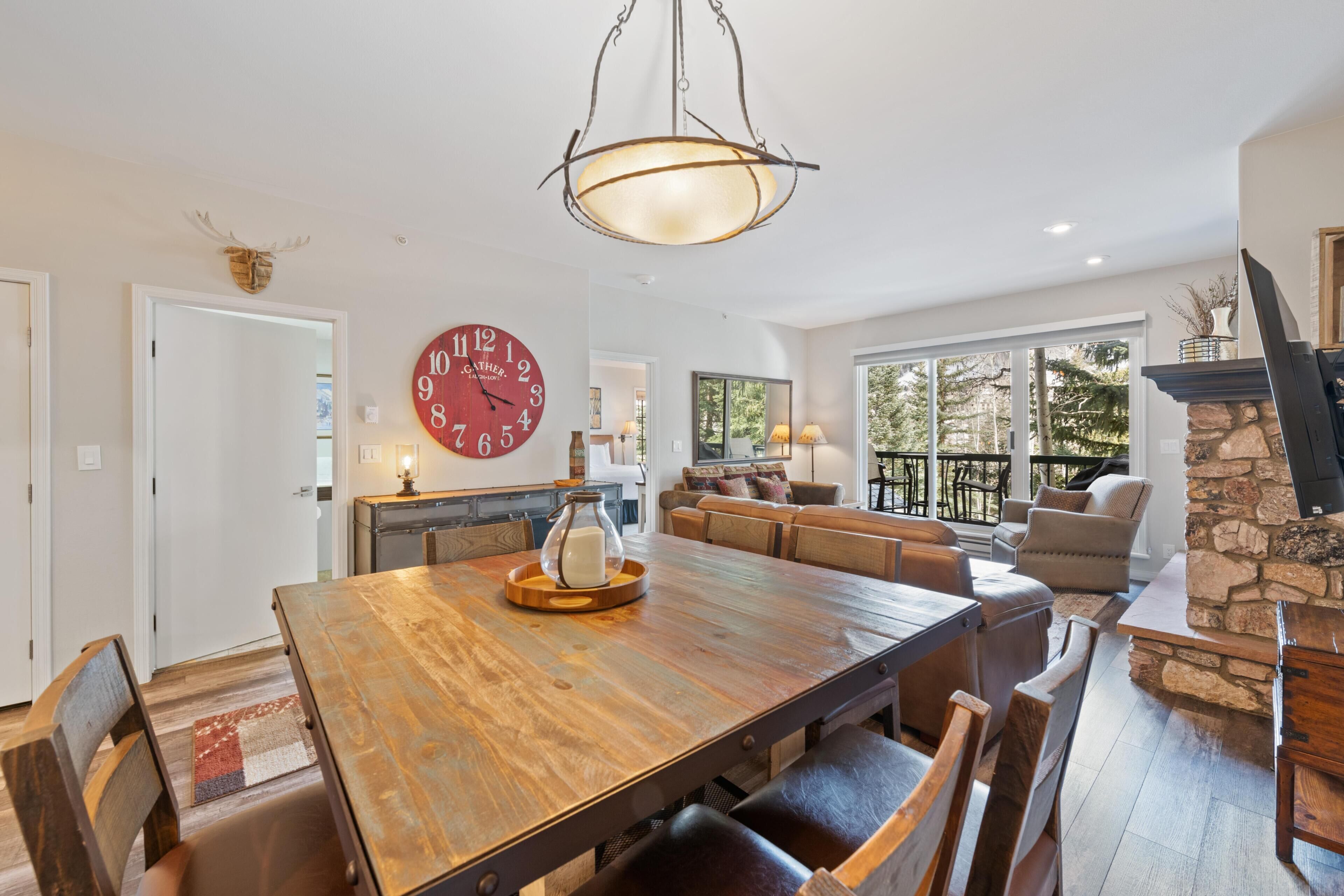 A spacious living area with a large wooden dining table, a stone fireplace, and a large clock on the wall.
