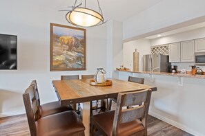A dining area with a wooden table and chairs, a painting on the wall, and a kitchen with white cabinets and appliances. - Borders Lodge U204 1BD Beaver Creek Ski-in, Ski-out Condo (Avon)