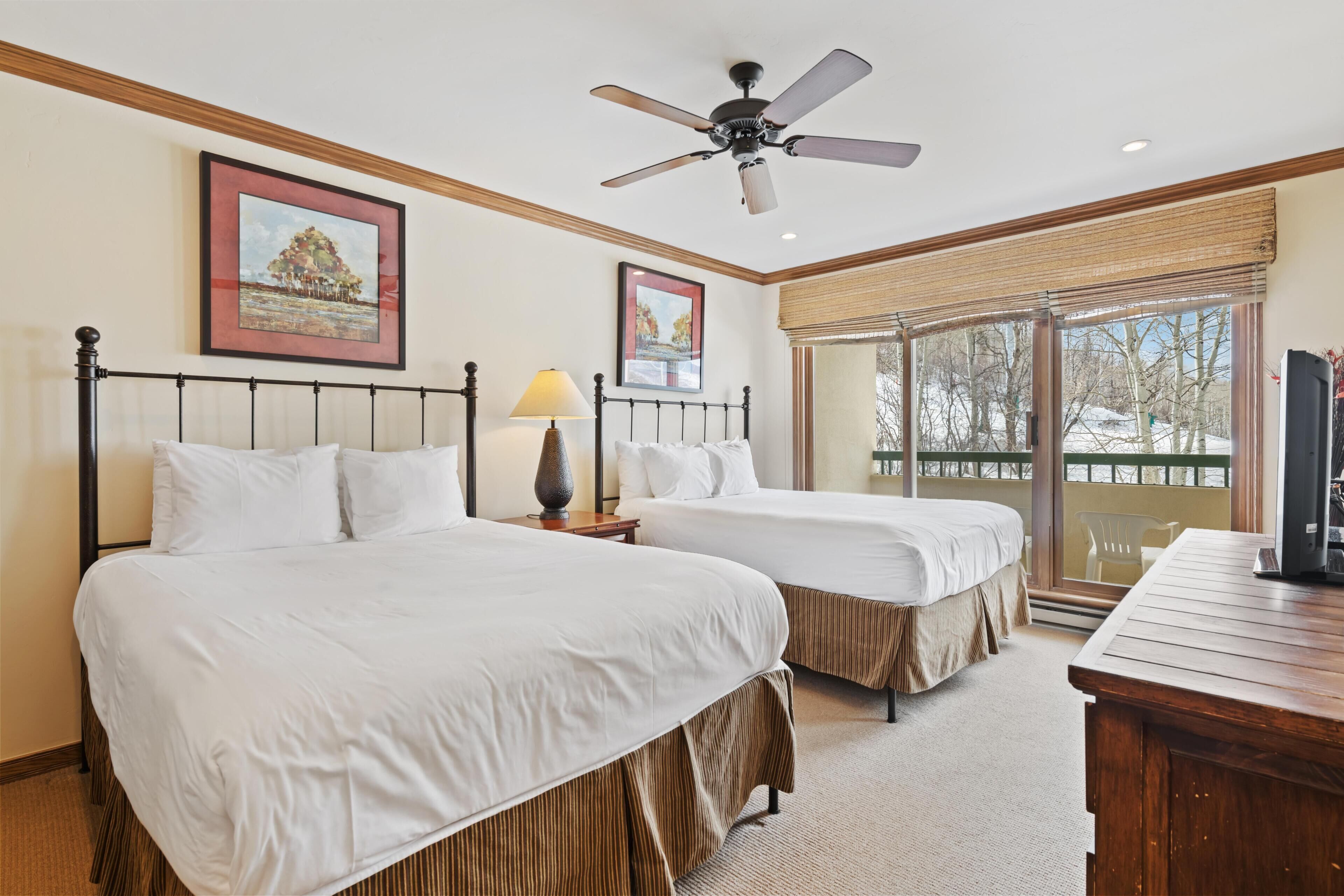 Une chambre d’hôtel avec deux lits, un ventilateur de plafond, une grande fenêtre donnant sur un beau paysage et un bureau en bois.