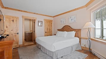 Un dormitorio con cabecera de madera, cama con ropa de cama blanca, lámpara de noche, ventana con vistas y puerta que da a un baño.