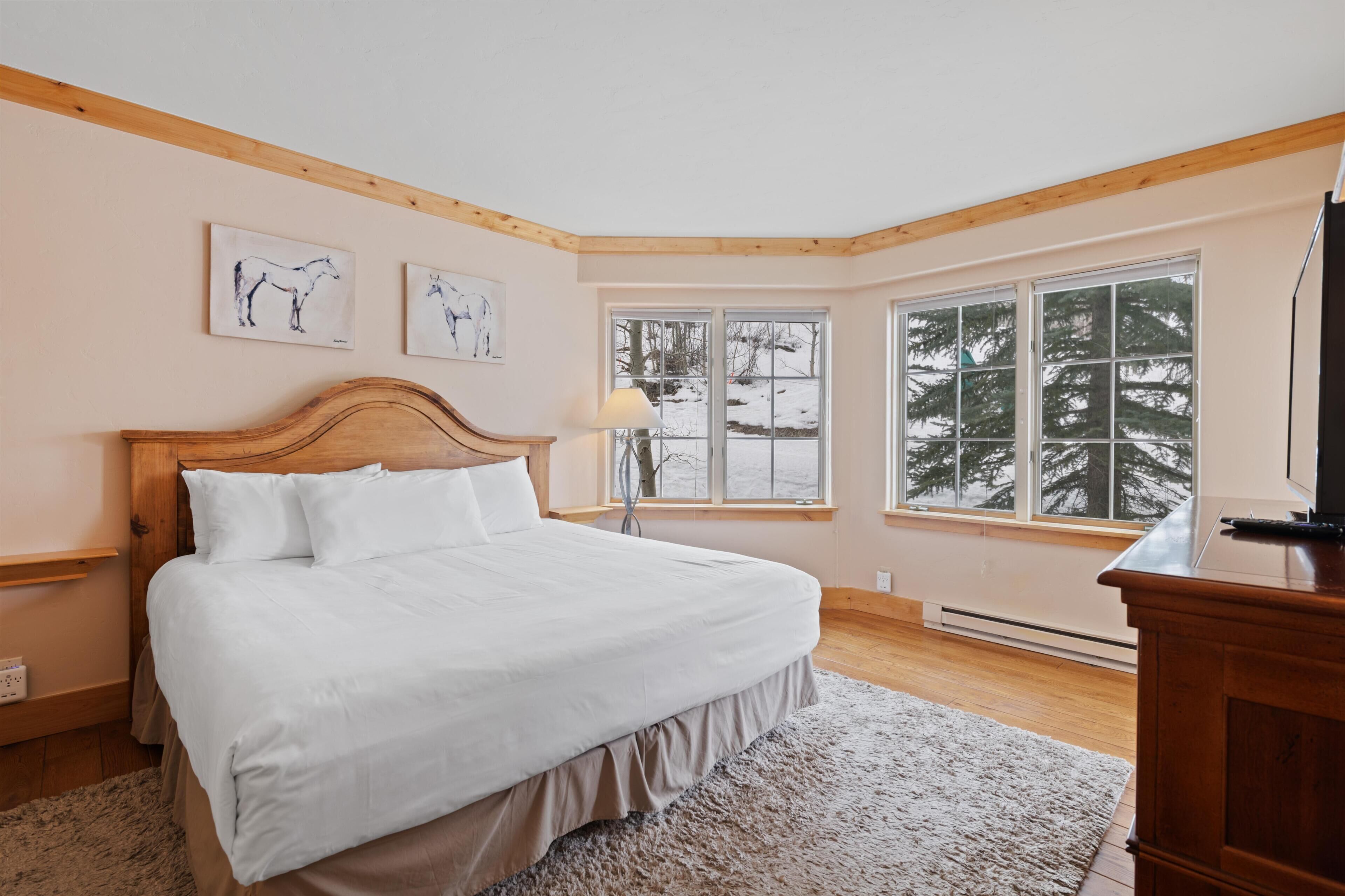 A bedroom with a wooden headboard, a large window with a view of trees, and a bed with white bedding.
