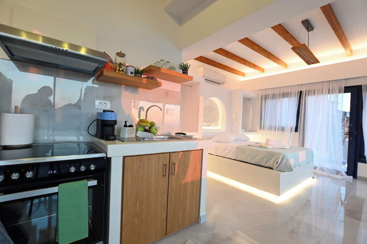 A modern kitchen with a stove, oven, and coffee maker, a bedside table with a bed, and a large window with sheer curtains.