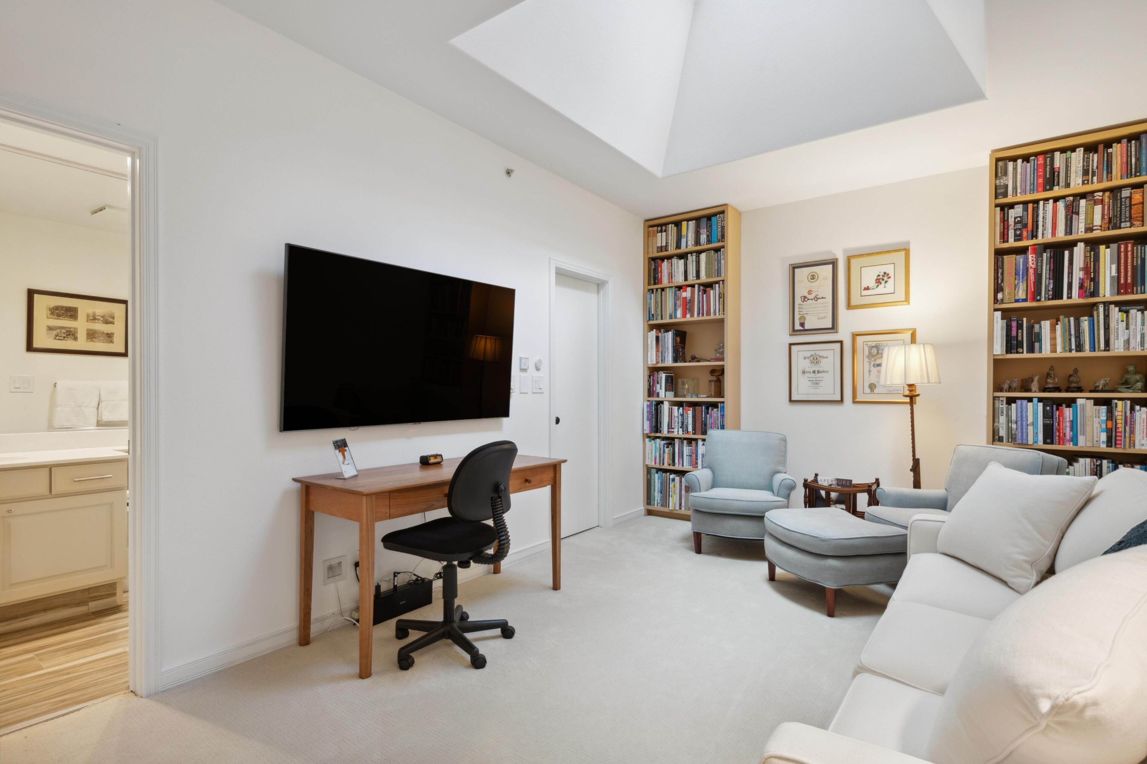 En rummelig opholdsstue med et fladskærms-TV, et skrivebord med stol, en sofa, en reol og en lampe.