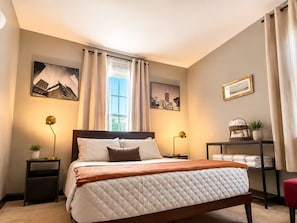 A hotel room with a bed, two bedside tables, a window with curtains, and wall art. - Historic Downtown Duplex - Fire Pit, Gym & Parking (Indianapolis)
