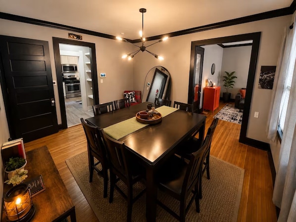 A dining area with a dark wooden table, multiple chairs, a central light fixture, and a kitchen in the background. - Historic Downtown Duplex - Fire Pit, Gym & Parking (Indianapolis)