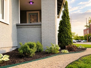 A building with a brick facade, a small porch, and a well-maintained garden with various plants. - Historic Downtown Duplex - Fire Pit, Gym & Parking (Indianapolis)