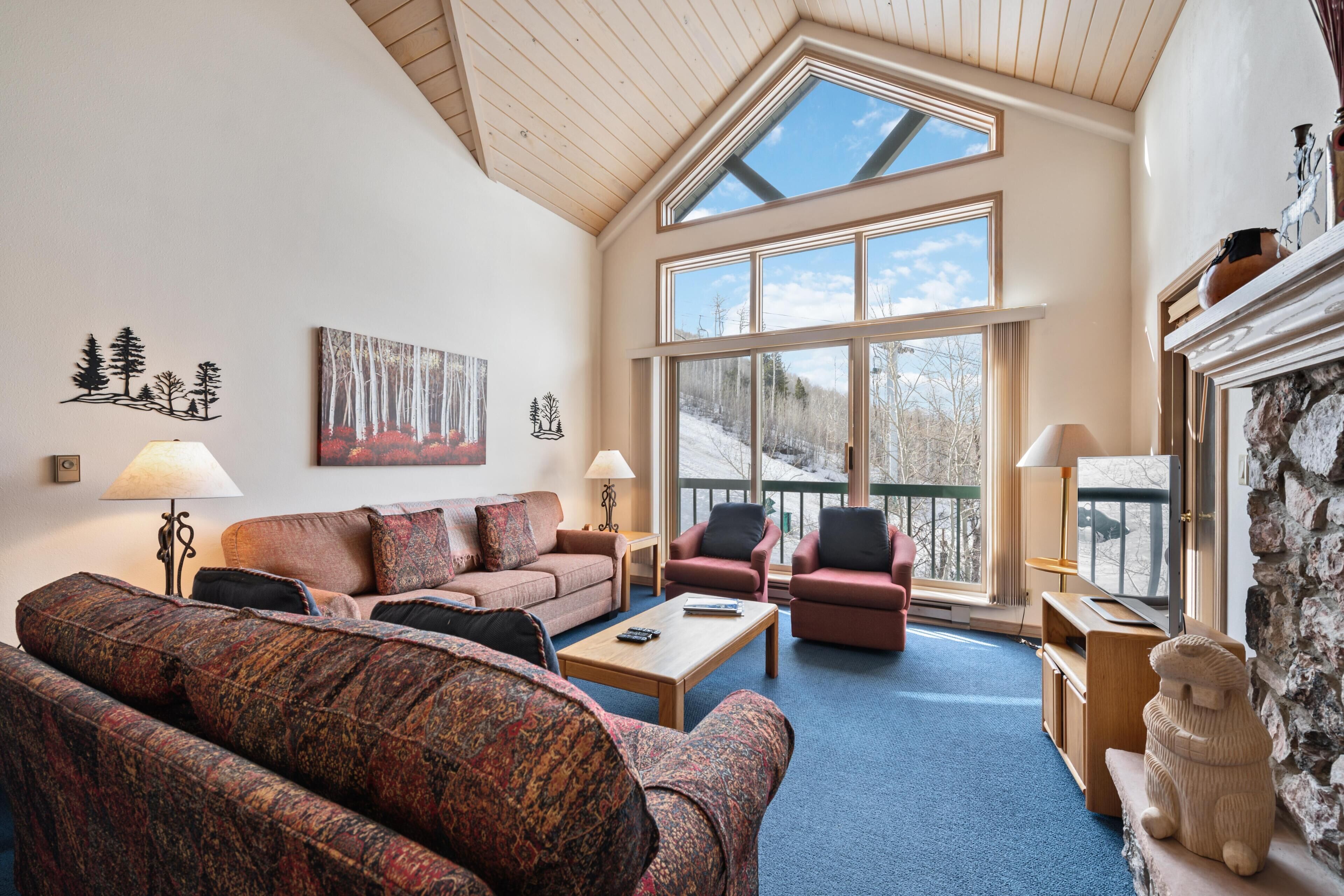 A cozy living room with a high ceiling, large windows, a fireplace, and a stone accent wall.