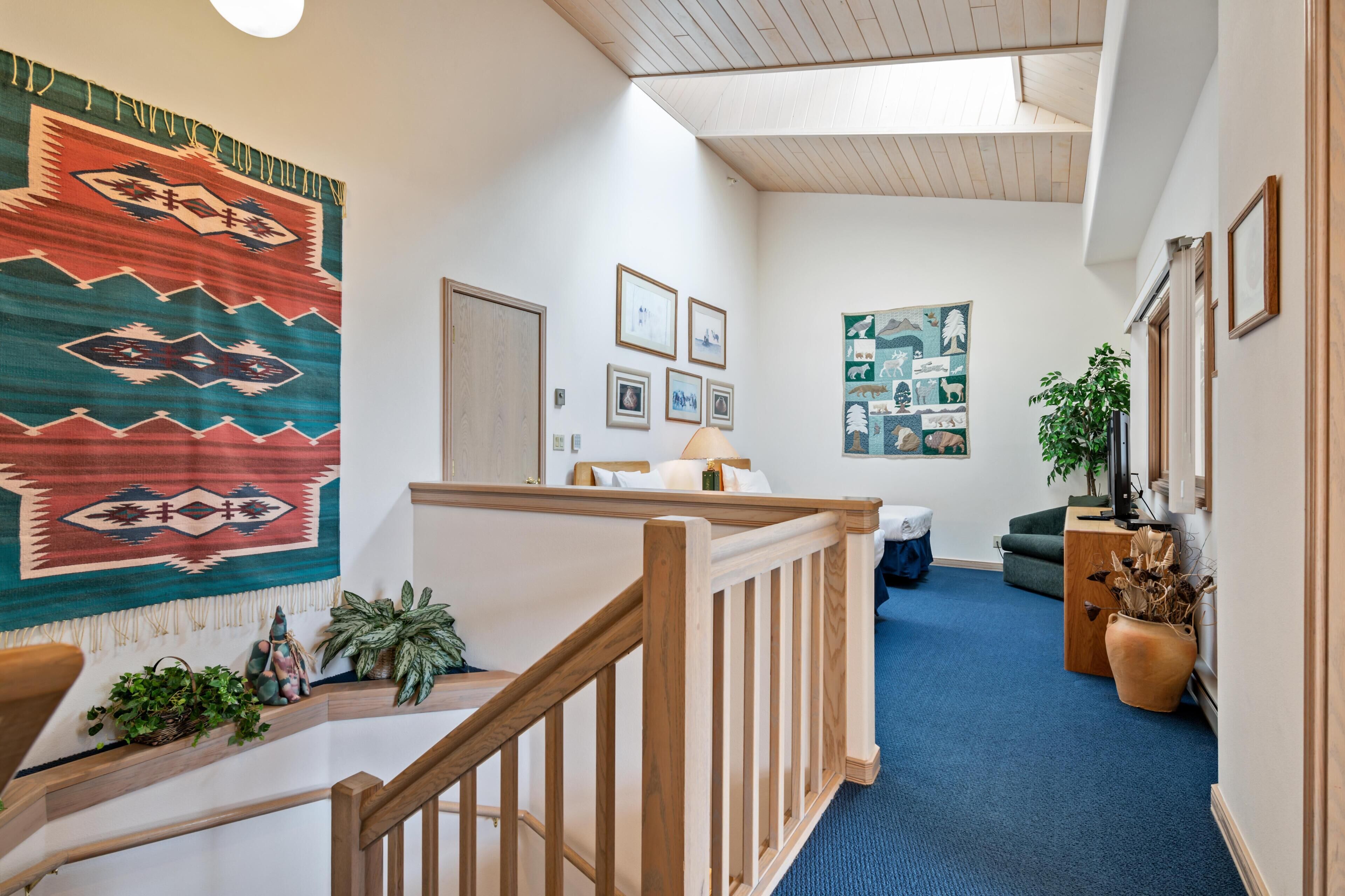 A hallway with a wooden railing, a blue carpet, a large Navajo rug, framed pictures, and a plant.