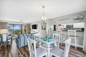 Ein modernes Wohnzimmer mit einem Glastisch, weißen Sesseln, einem Sofa mit blauen und weißen Streifen, einer Küchenzeile mit weißen Schränken und Blick auf den Ozean.