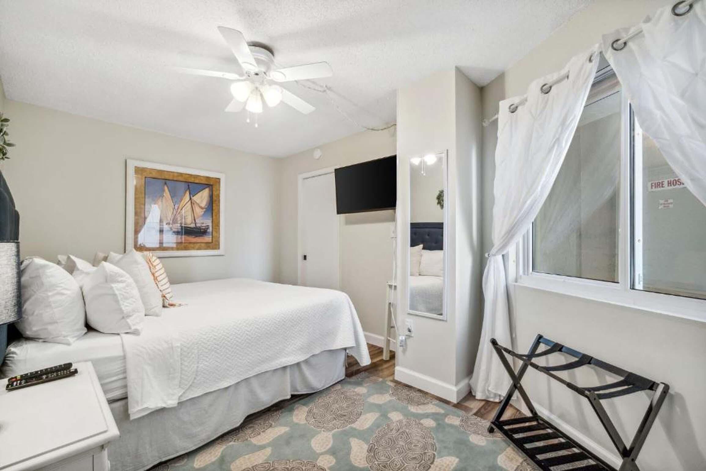 Une chambre à coucher comprenant un lit, un ventilateur de plafond, une fenêtre avec des rideaux, une table de chevet et un téléviseur fixé au mur.