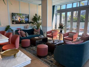 A modern hotel lobby with a sofa set, armchairs, ottomans, a coffee table, and a large window offering a view of the outdoors. - Luxury Designer Retreat in Wynwood / Skyline. 2BR. Pool & Bar (Miami)