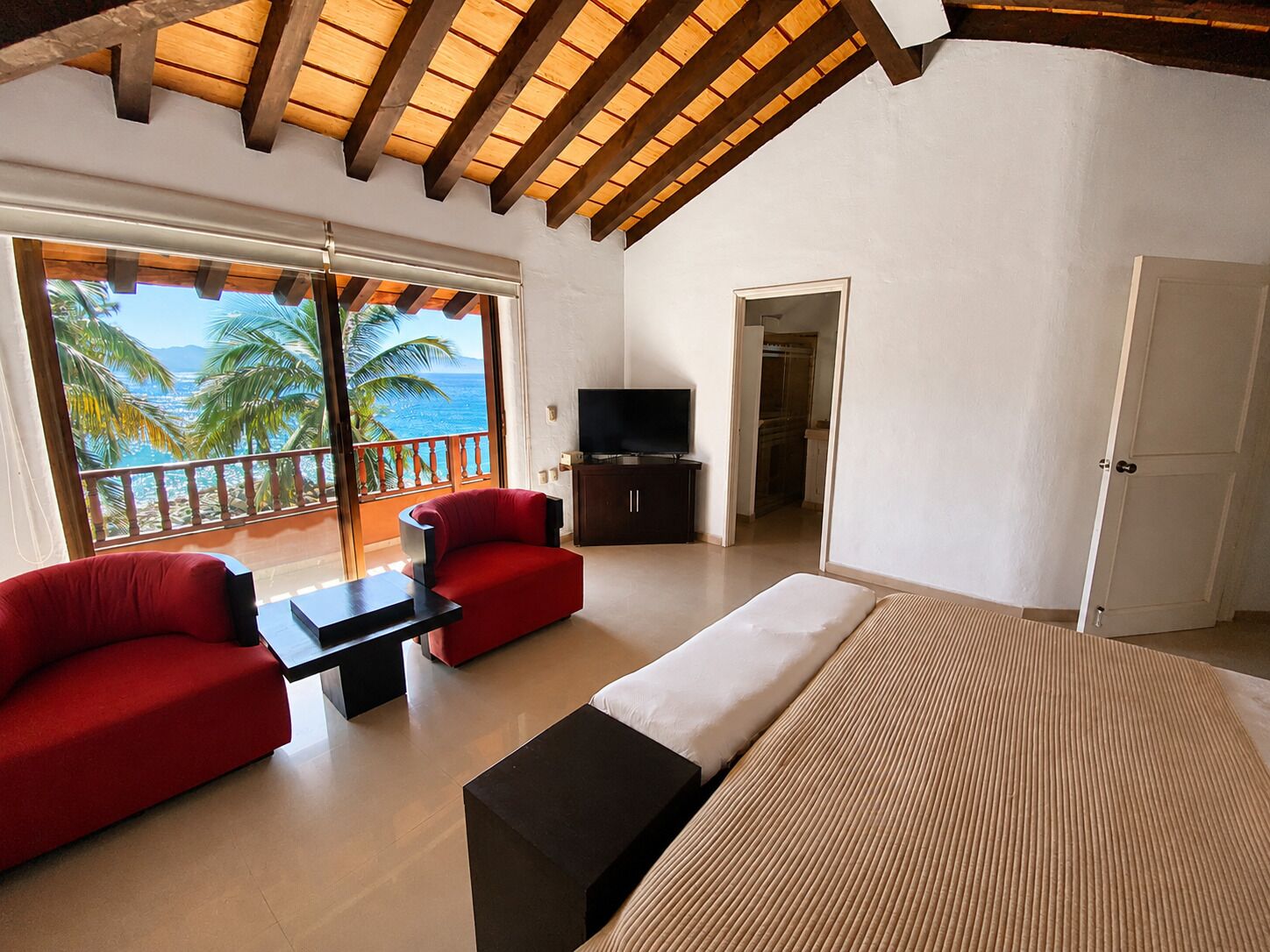 A bedroom with a bed, a TV, a balcony with a view of palm trees and the sea, and a red sofa.