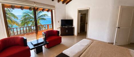 A bedroom with a bed, a TV, a balcony with a view of palm trees and the sea, and a red sofa.