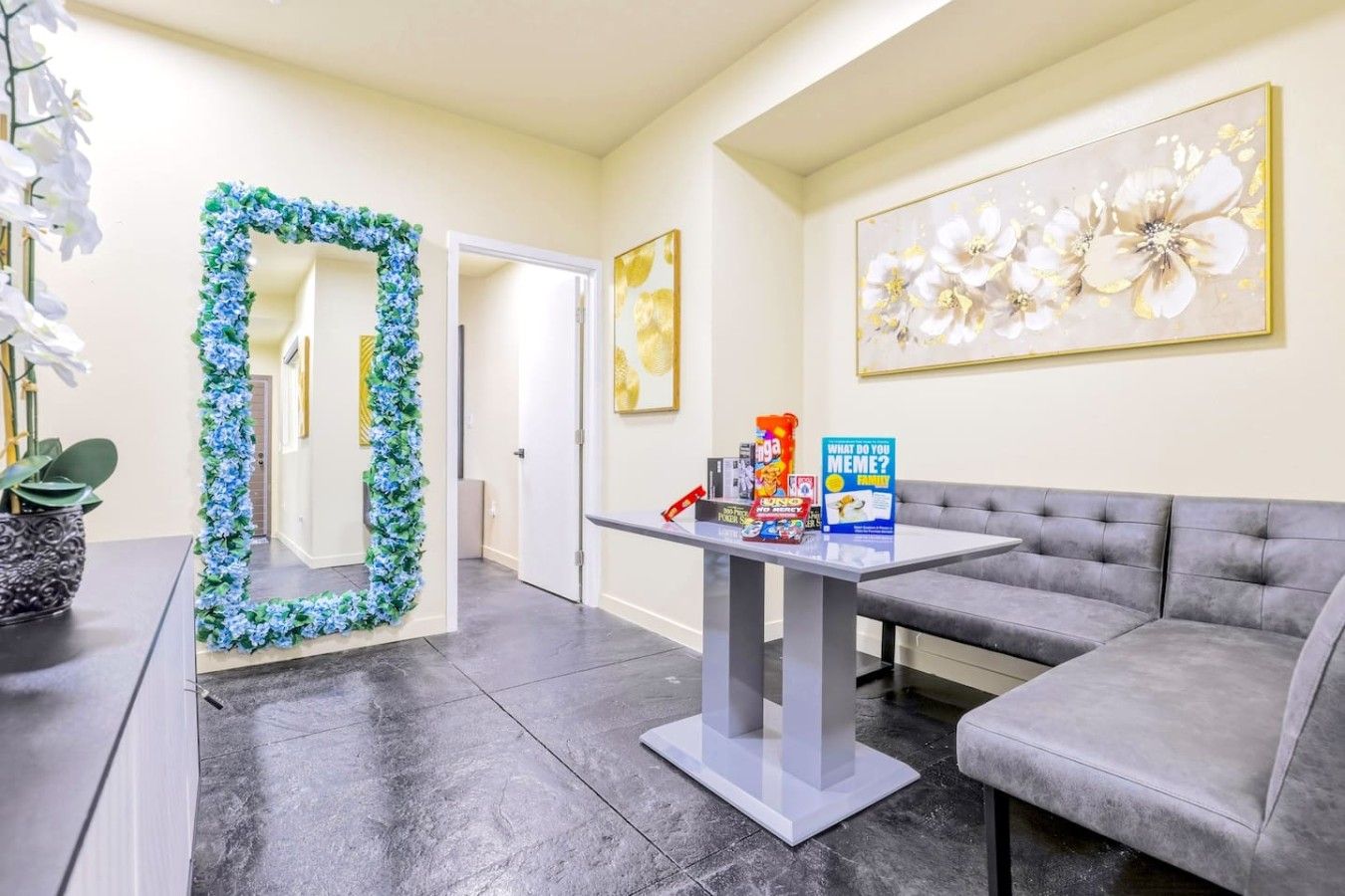 Een moderne woonkamer met een grijze bank, een salontafel met boeken en een gezelschapsspel, een decoratieve spiegel en een bloemig kunstwerk aan de muur.