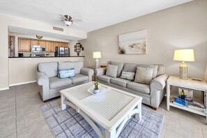 Ein Wohnzimmer mit zwei grauen Sofas, einem weißen Couchtisch, einer Küche im Hintergrund und einem Deckenventilator.