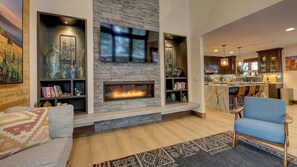 A living room with a fireplace, bookshelves, a sofa, and a dining area with a kitchen in the background.