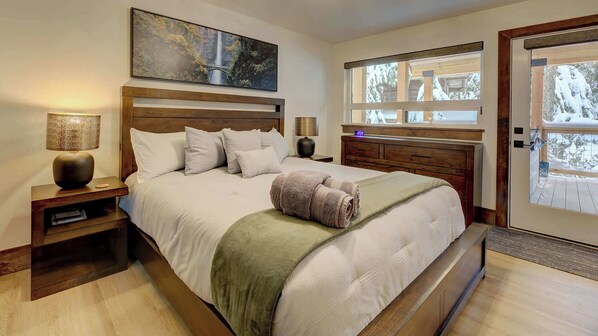 A bedroom with a large bed, wooden headboard, bedside tables, a lamp, a painting, and a window with a view of trees.
