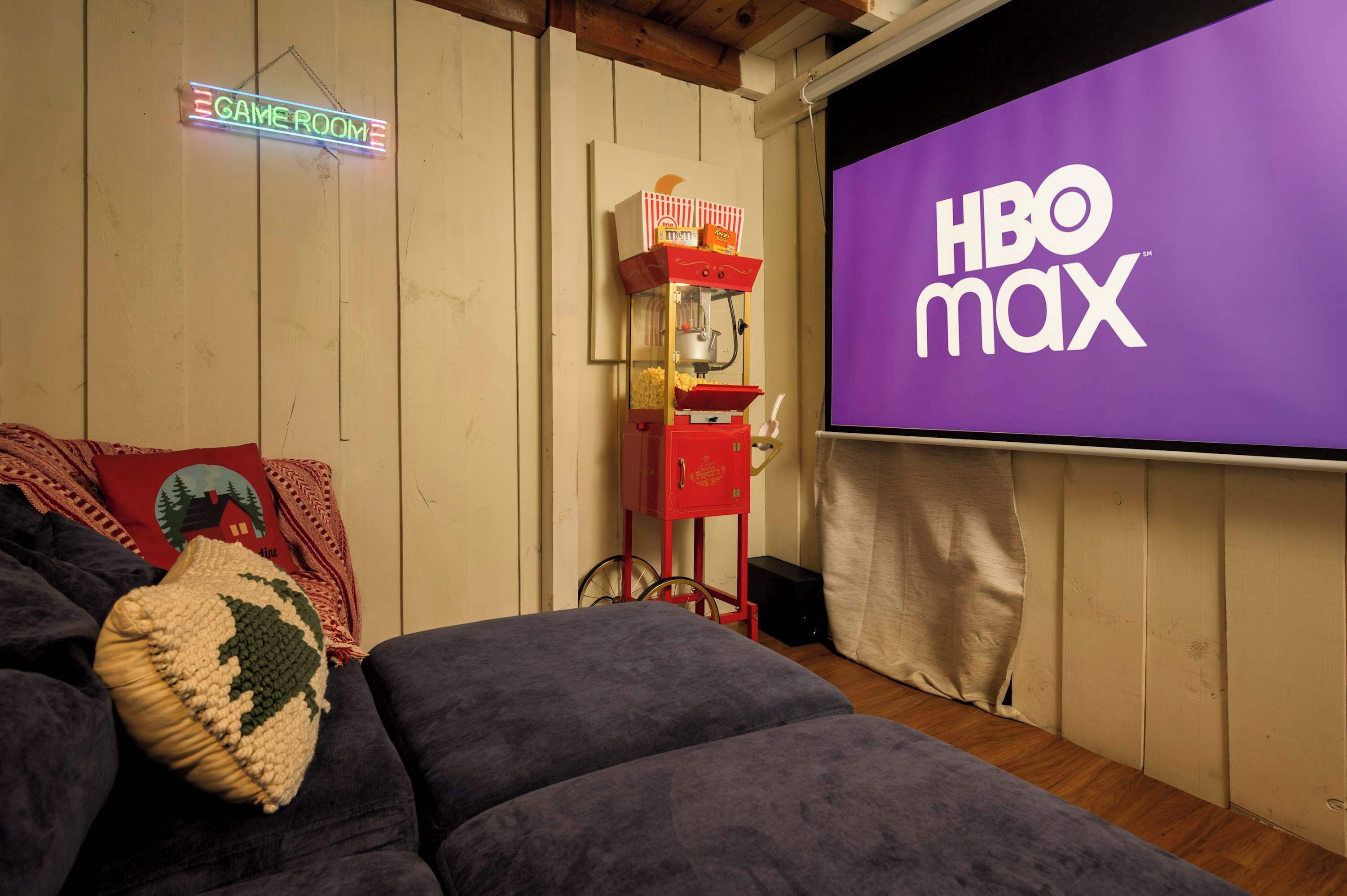 A room with a couch, a neon sign reading 'GAMEROOM,' a popcorn machine, and a large screen displaying 'HBO max.'