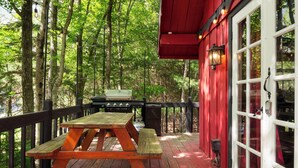 Una terraza de madera con una mesa de picnic y una parrilla, rodeada de árboles.