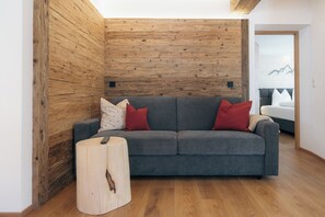 Ein modernes Wohnzimmer mit grauem Sofa, Holzvertäfelung, einem runden Holzhocker und einem Bett mit Bergdekor im Hintergrund.