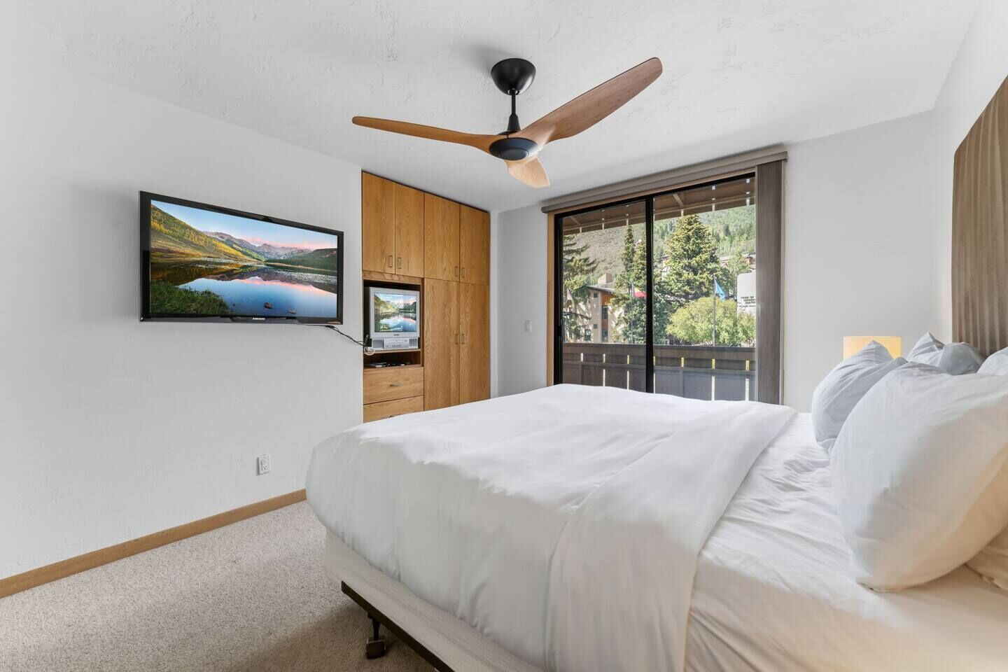 Un dormitorio con cama, ventilador de techo, TV montada en la pared y ventana con vista a vegetación.