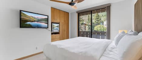Un dormitorio con cama, ventilador de techo, TV montada en la pared y ventana con vista a vegetación.