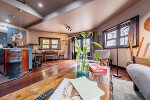 A living room with a wooden floor, a sofa, a coffee table with a book and a vase, a dining area with a table and chairs, and a kitchen area with a counter and appliances. - 
Beachfront  Apartment – Wake Up to Caribbean Sea Views (Hopkins Village)