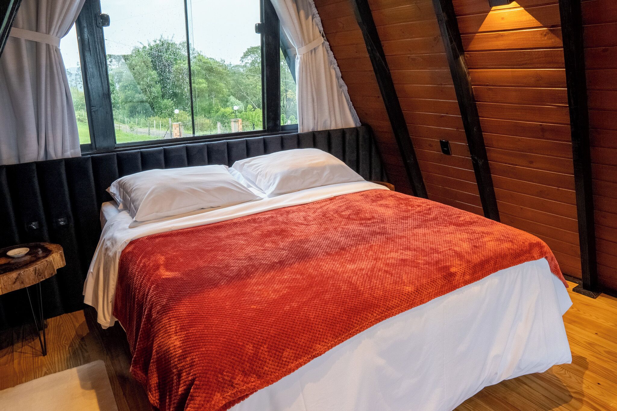 A room with a bed, a window with curtains, a wooden floor, and a small wooden table.