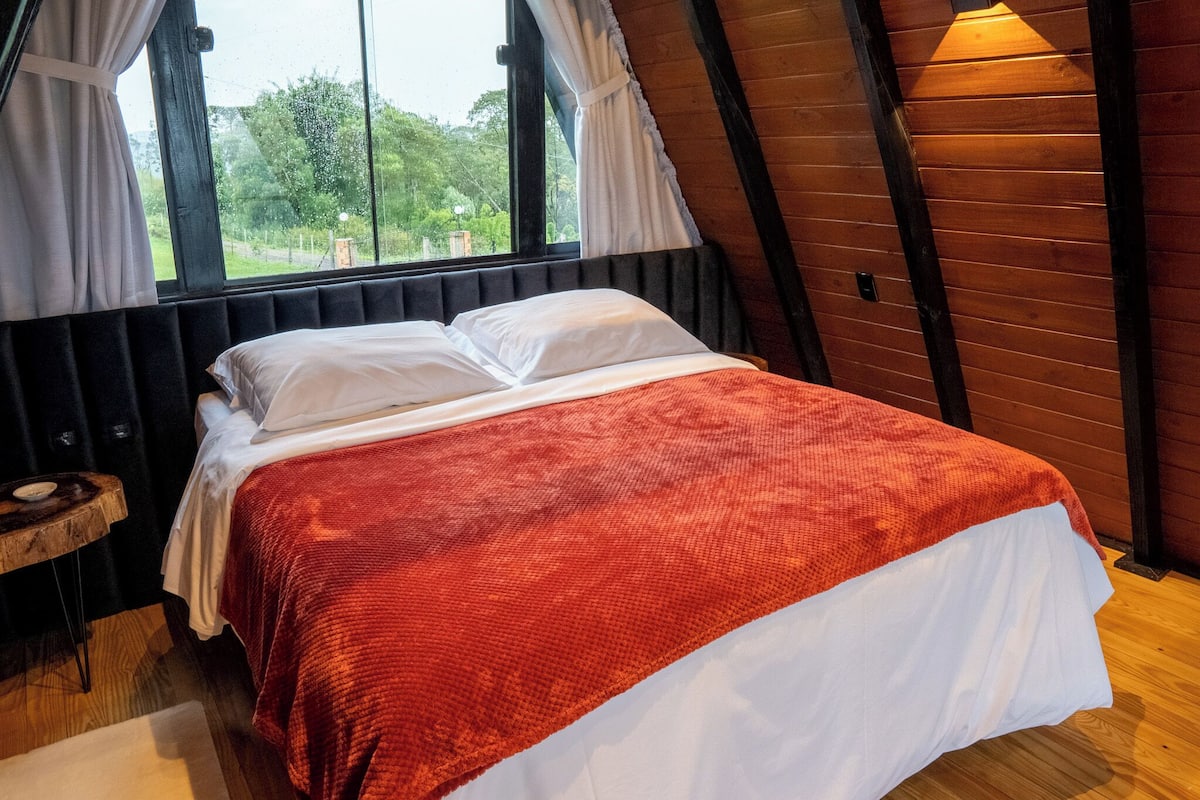 Una habitación con cama, ventana con cortinas, suelo de madera y una pequeña mesa de madera.