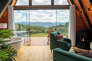 A cozy room with a large window, a green armchair, a white bathtub, and a wood-burning stove. - Altitude 1140 Chalés (Águas Brancas)