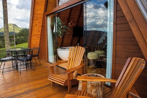 A wooden deck with a table and chairs, a large white bathtub, and a glass door leading to a balcony with a view of a mountain. - Altitude 1140 Chalés (Águas Brancas)