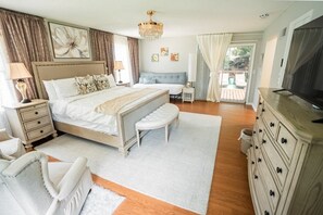 A bedroom with a bed, bedside tables, a bench, a dresser, a television, and a chandelier. - 5 Mins to Arrowhead | 3BR KC Retreat | 3 Ensuites, Sauna & Theater (Independence)
