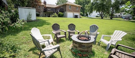 Patio trasero con un brasero, cuatro sillas Adirondack y un césped.