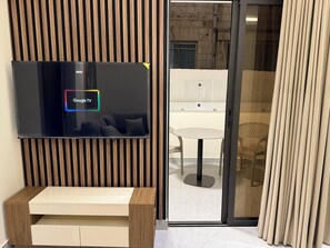 A modern hotel room with a flat-screen TV mounted on a wooden panel wall, a glass door leading to a balcony, and a wooden bench. - 002 JRE Apartment With Stunning Boulevard Veiw (Amman)