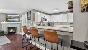A modern kitchen with a fireplace, bar stools, and a large window. - Alpine Escape | Pool, Hot Tub & Mountain Access (Edwards)