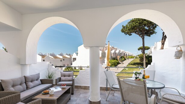 Un espacio de estar al aire libre cubierto con sofá, sillas y una mesa, con vista a una hilera de edificios blancos y un árbol.