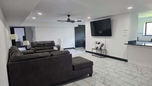 Un soggiorno moderno con un ampio divano scuro, una TV a schermo piatto montata a parete, un ventilatore a soffitto e un pavimento piastrellato.