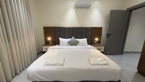 A modern hotel room with a large bed, two bedside tables with lamps, a ceiling air conditioner, and a door. - 003jre Apartment With Stunning Boulevar (Amman)