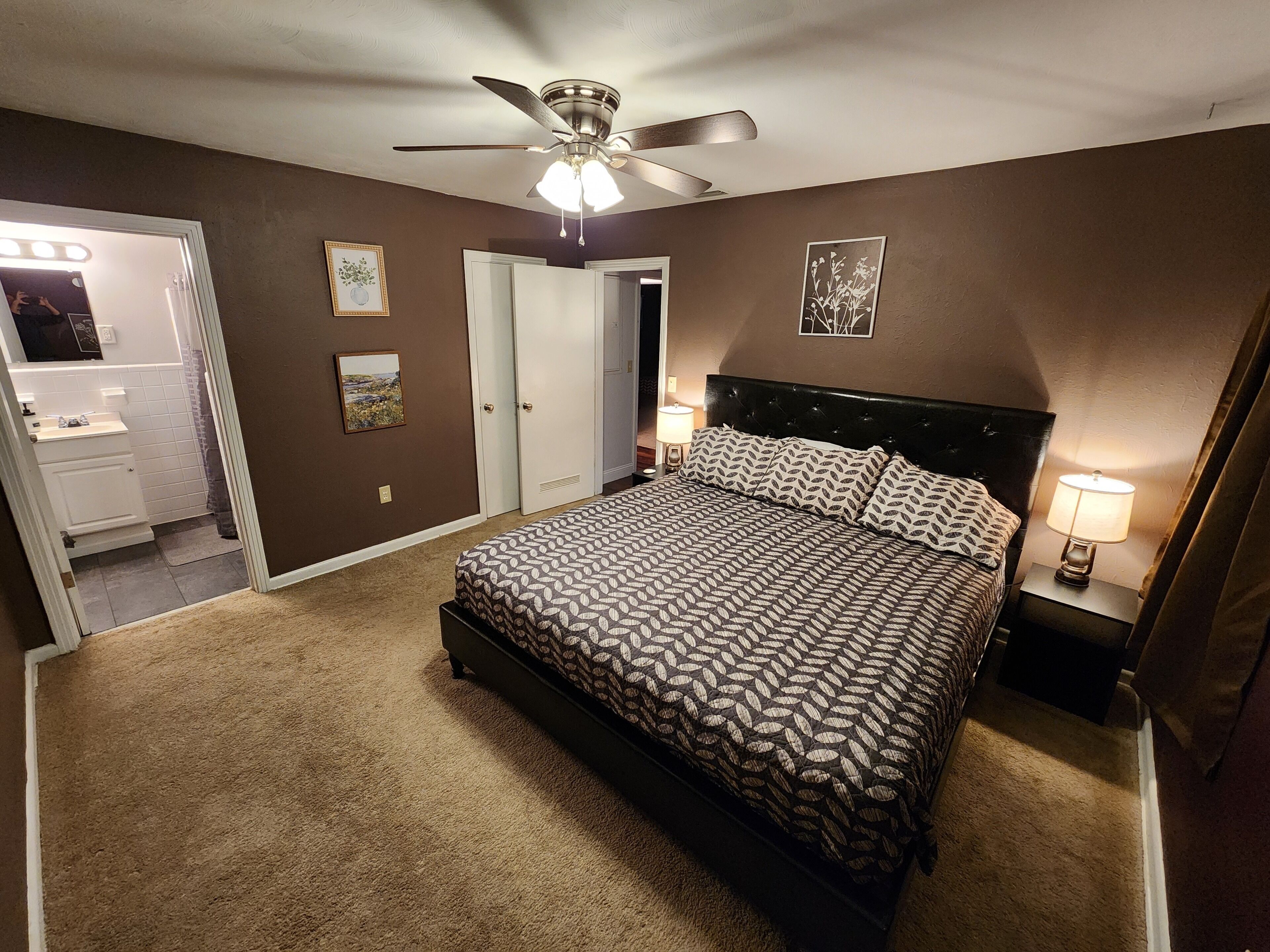Un dormitorio con cama, ventilador de techo, mesita de noche con lámpara y puerta que da a un baño.