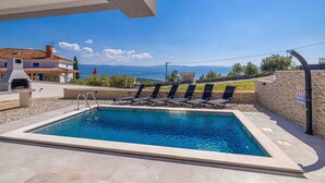 En swimmingpool med liggestole, grillområde og udsigt til et bjergrigt landskab.