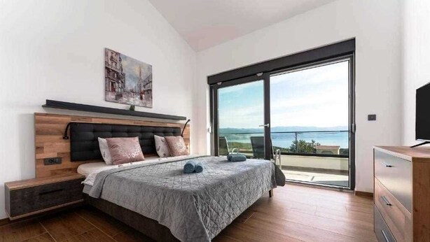 Et soveværelse med en stor seng, et træhovedgærde, et maleri, en skydedør til en balkon med udsigt og et tv-møbel.