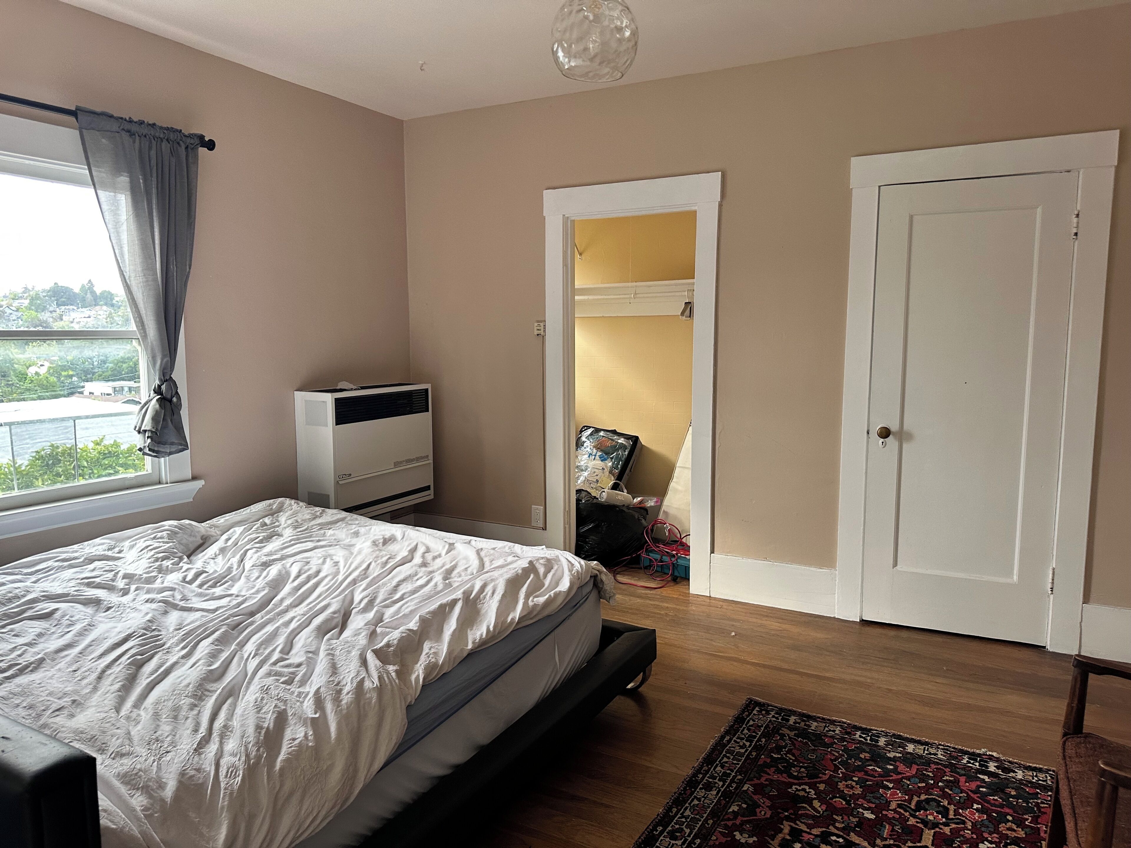 A bedroom with a bed, an air conditioner, a window with curtains, a door, and a view of the outside.