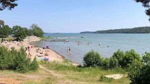 En strand med folk som badar och solar, en brygga och träd i bakgrunden.